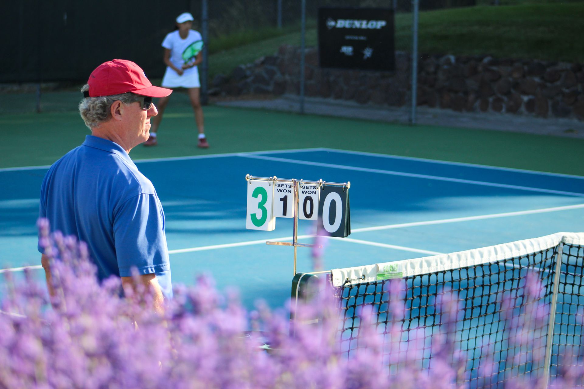 Tennis official beside tennis court scoreboard, with blurred flowers in foreground and tennis player in background.