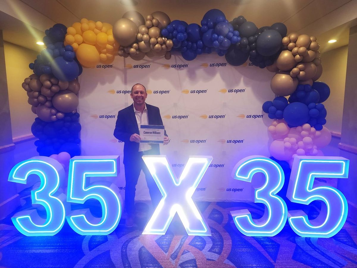 Man standing in front of a US Open backdrop with illuminated "35 x 35" sign and balloon arch in shades of blue, gold, and purple.