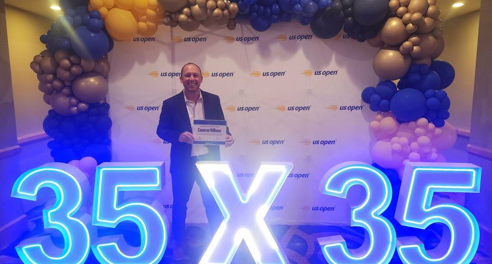 Man standing in front of a US Open backdrop with illuminated "35 x 35" sign and balloon arch in shades of blue, gold, and purple.