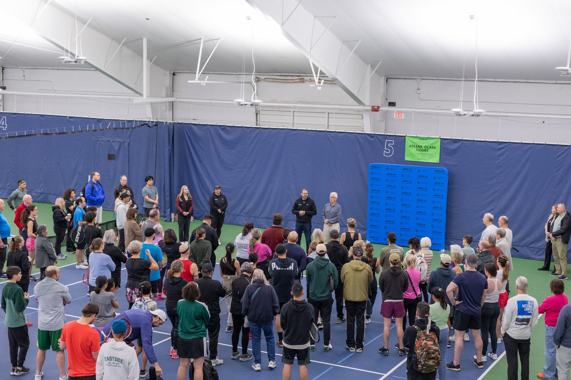 A group of people gathered on indoor tennis courts, listening to speakers in front of a backdrop, with a "Arlene-Clark Court" sign visible.