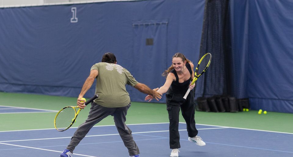 Two people on an indoor tennis court playfully high-five while holding rackets, with tennis balls scattered around.