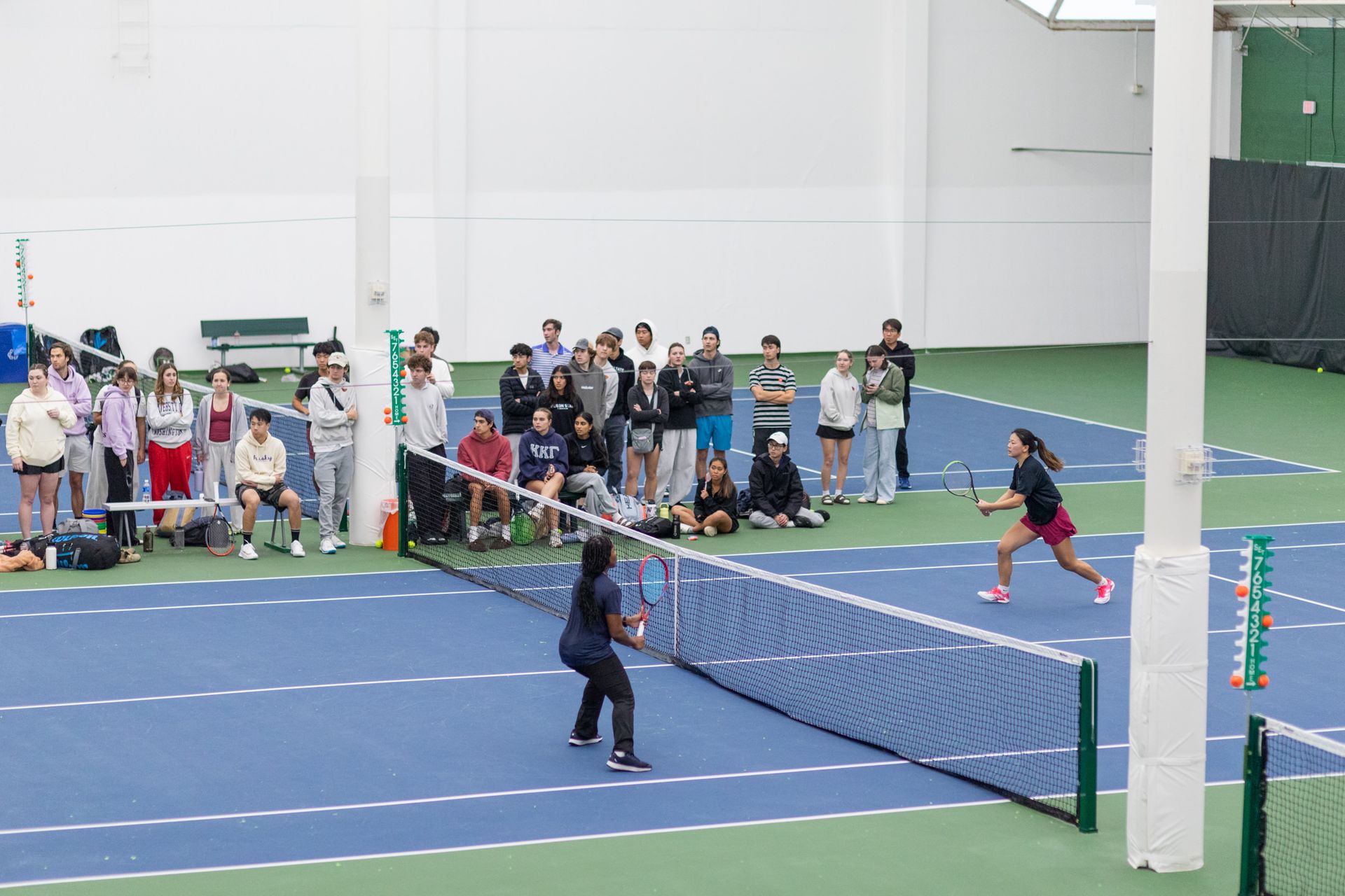 Two people play tennis indoors with a crowd watching intently from the sideline on a blue and green court.