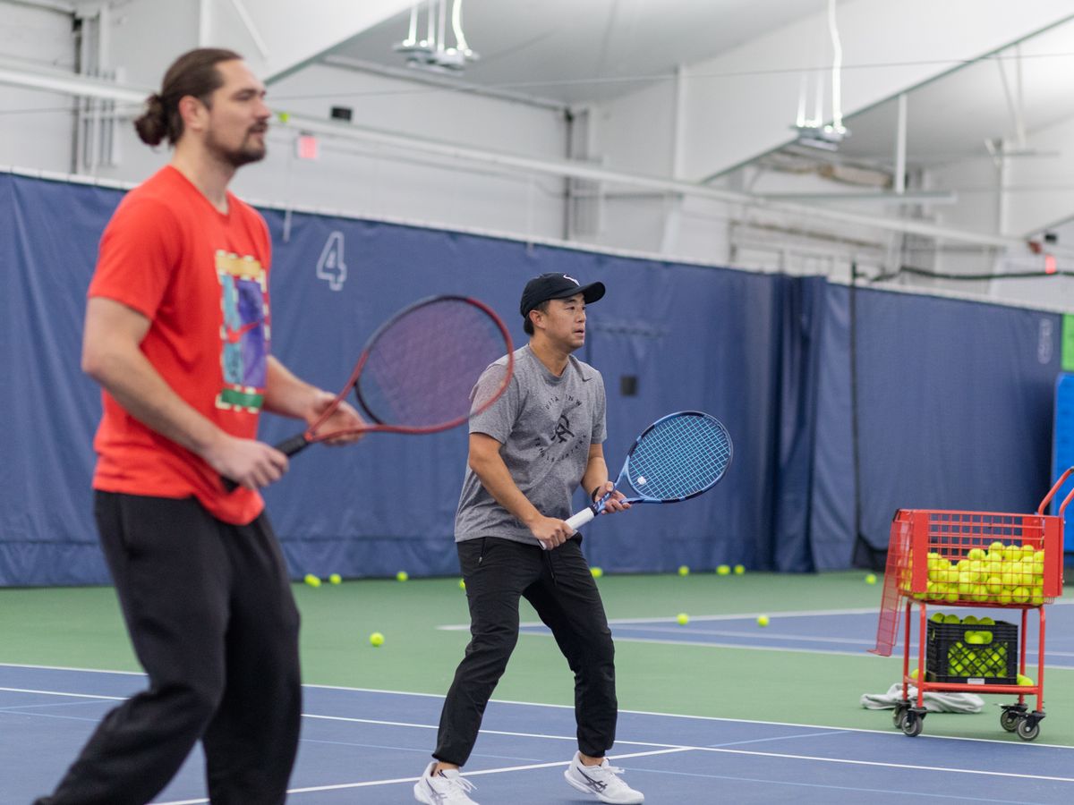 Two men playing tennis indoors with a practice basket filled with tennis balls nearby.