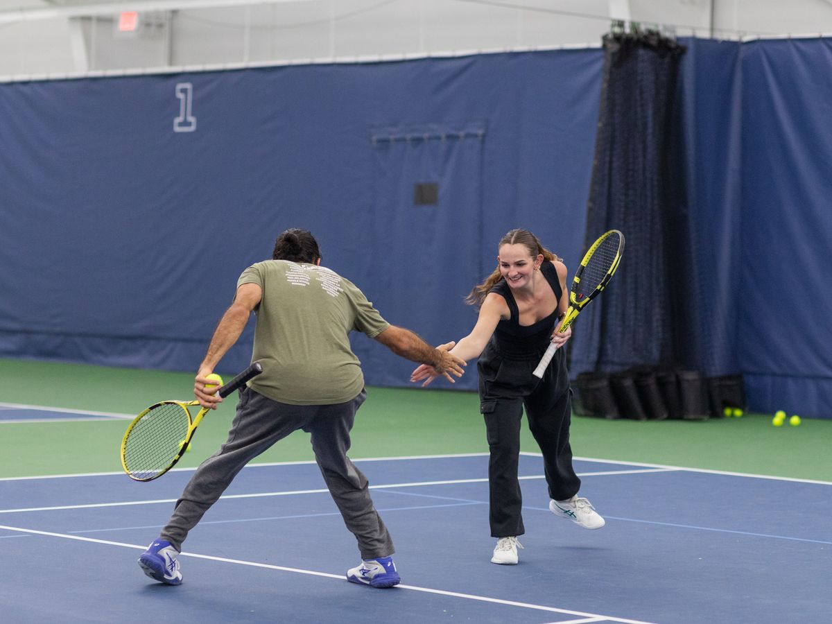 Two tennis players, a man and a woman, high-five on an indoor court, both holding rackets and smiling.