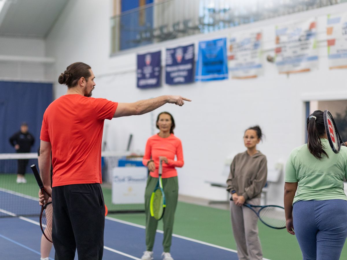 Tennis coach in red shirt instructs three female players holding rackets on an indoor court.