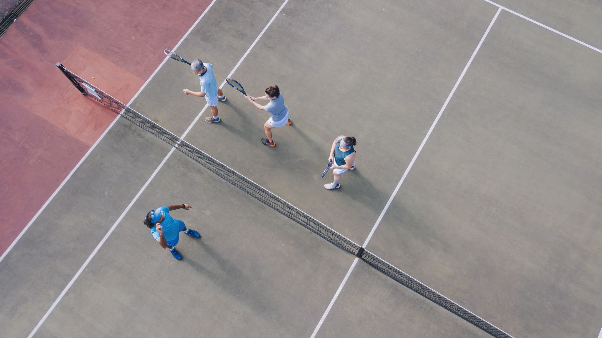 Aerial view of four people with tennis rackets on a court, positioned around the net, preparing for a game.
