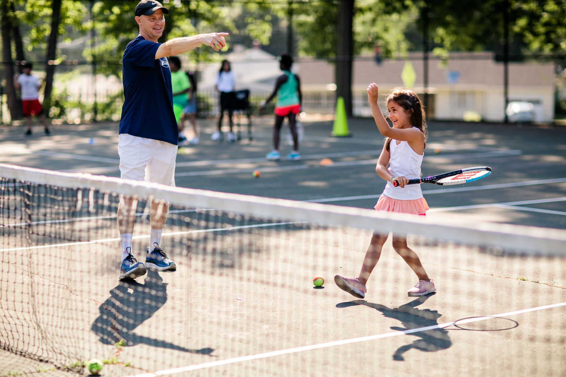 A tennis coach guides a young girl on a court. She’s smiling, holding a racket, surrounded by tennis balls and distant players.