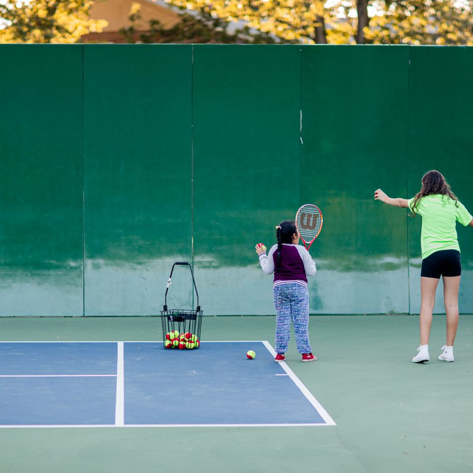 Two people practice tennis swings on a court against a green wall, with a basket of tennis balls nearby.