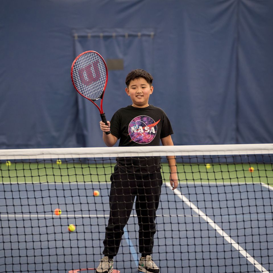 Boy in a NASA shirt holding a tennis racket on an indoor court, with tennis balls scattered on the blue surface.