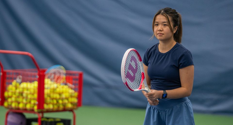 A woman in sports attire holds a tennis racket on an indoor court, with a cart of tennis balls nearby.