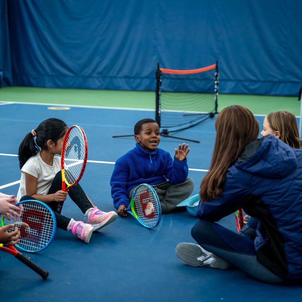 Children sitting on an indoor tennis court, holding rackets, engaged with their instructor. A trolley with tennis balls is visible nearby.