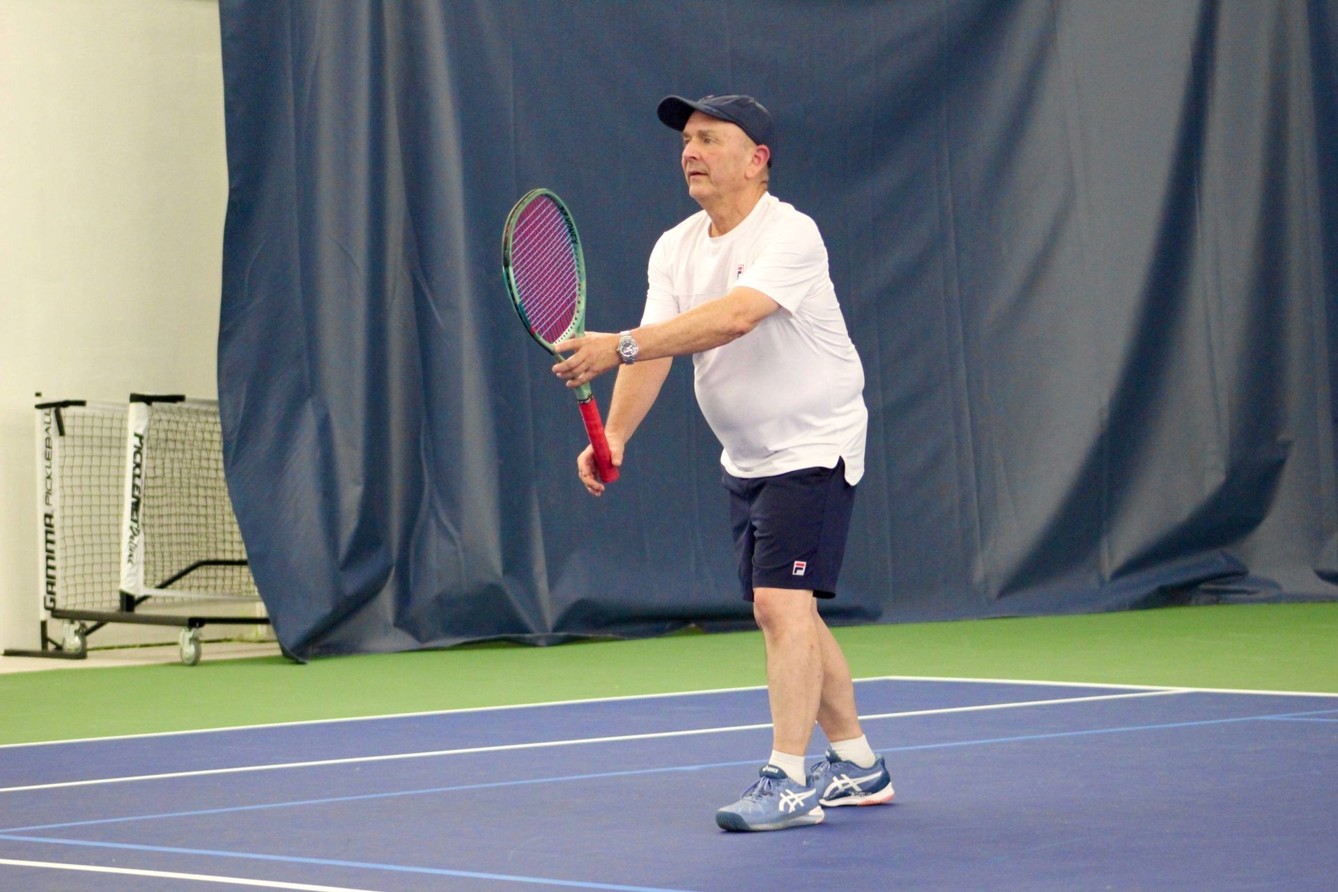 Older man playing tennis indoors, wearing a white shirt, navy shorts, and a cap, holding a red and purple racket on a blue court.