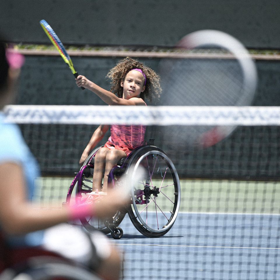 Two young girls in wheelchairs playing tennis, one hitting the ball, the other watching from across the court.