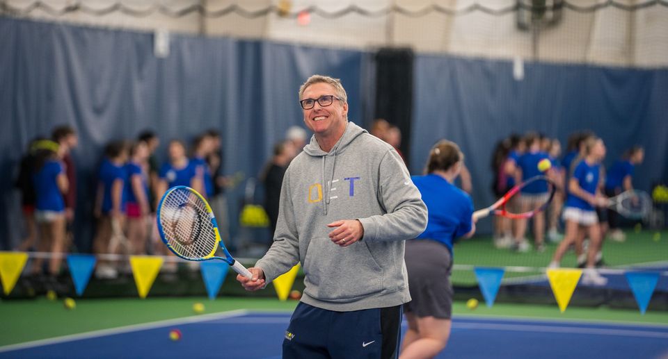 A smiling man in a grey hoodie holds a tennis racket on an indoor court with a group of people in the background.