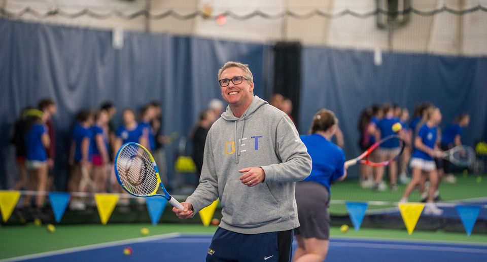 A smiling man in a grey hoodie holds a tennis racket on an indoor court with a group of people in the background.