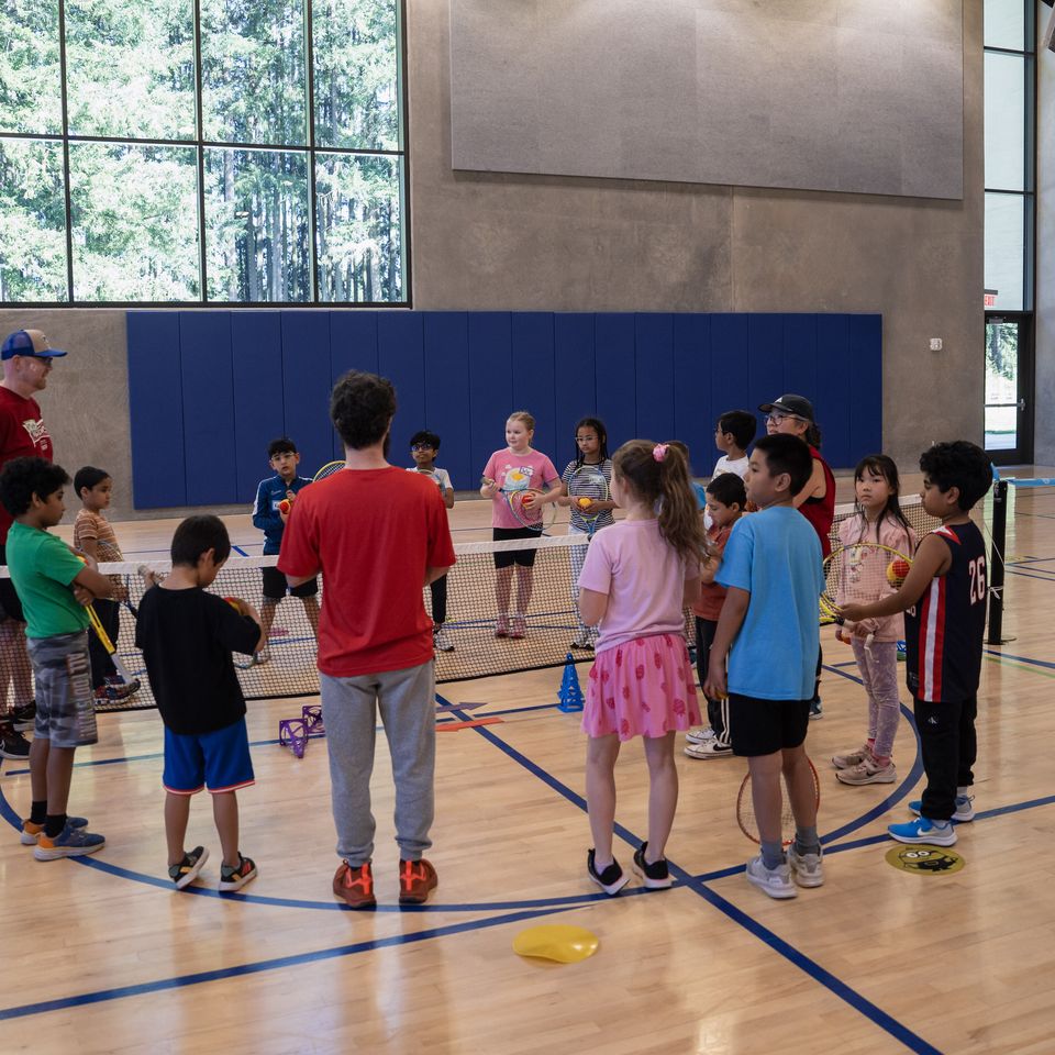 Children and instructors gather in a gym for a tennis lesson, with rackets and colorful cones on the court.