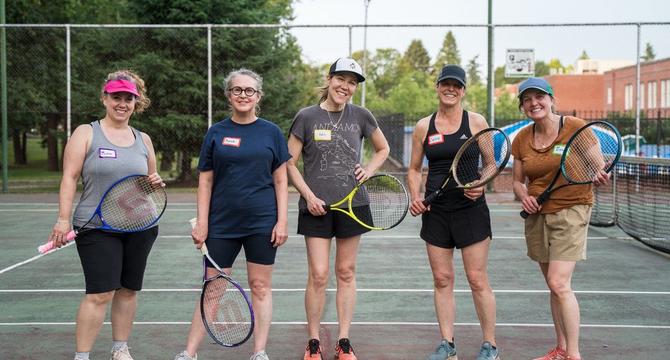 Five women smiling on a tennis court, holding rackets, wearing casual sportswear and name tags, with trees and buildings in the background.