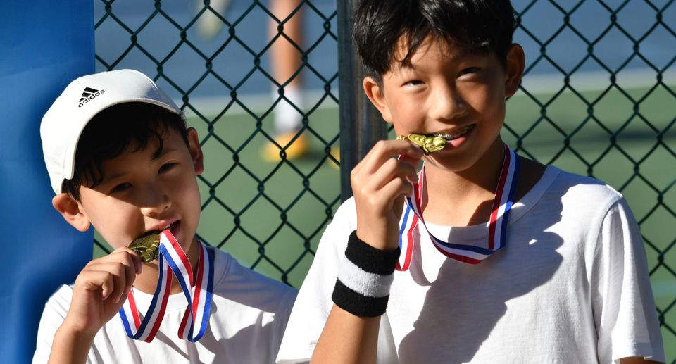 Two boys in white shirts, wearing medals, pose by a tennis court fence, playfully biting their medals and smiling.