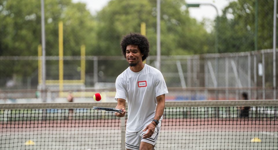 A man in a white shirt plays tennis outdoors, focusing on a red and yellow ball above his racket. A net and trees are in the background.