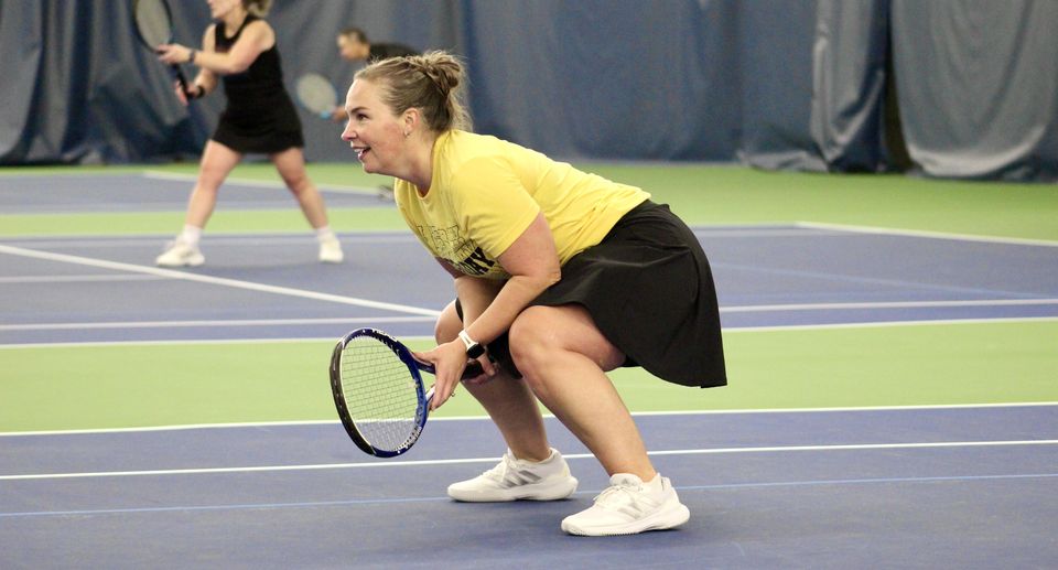 Two women playing doubles tennis indoors, one in yellow ready to receive, the other in black behind her in position.