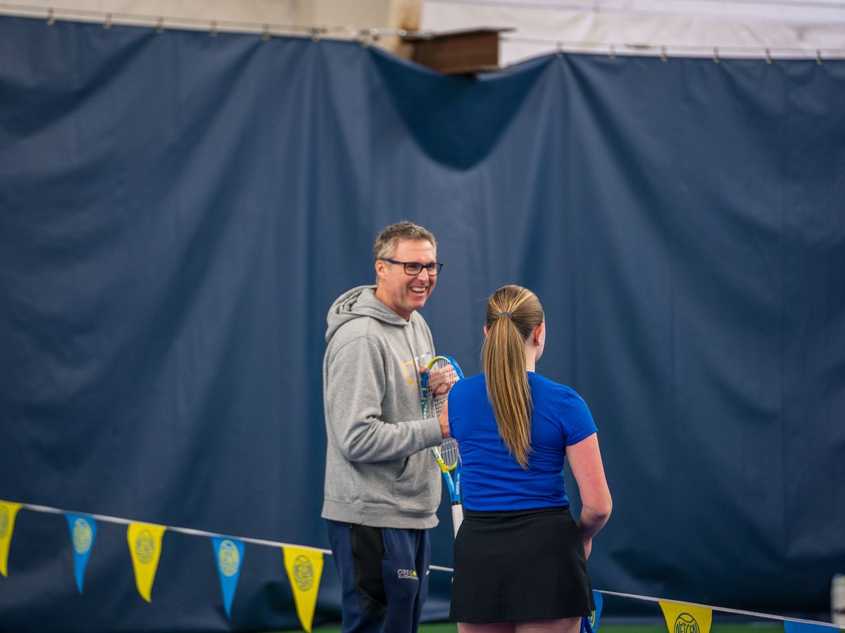 A man in a grey hoodie and a woman in a blue shirt converse and smile at an indoor tennis court.