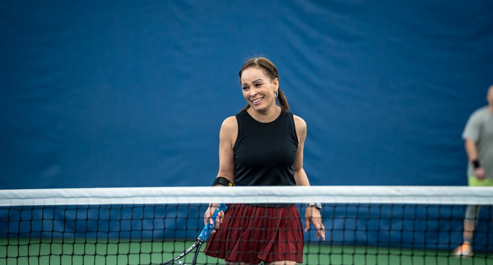 Woman smiling on an indoor tennis court, holding a racket near the net, wearing a black top and red skirt.