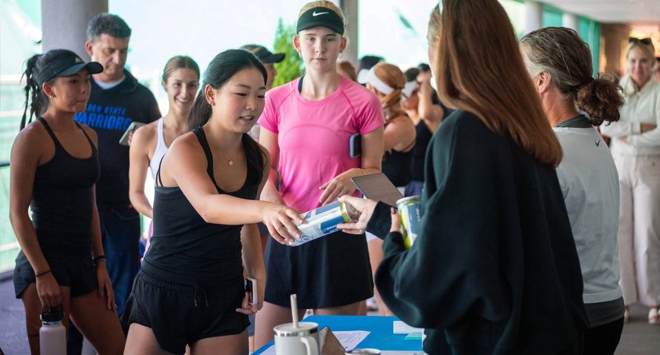People in athletic wear gather at a table, one woman handing out items, possibly at a sports event or registration area.