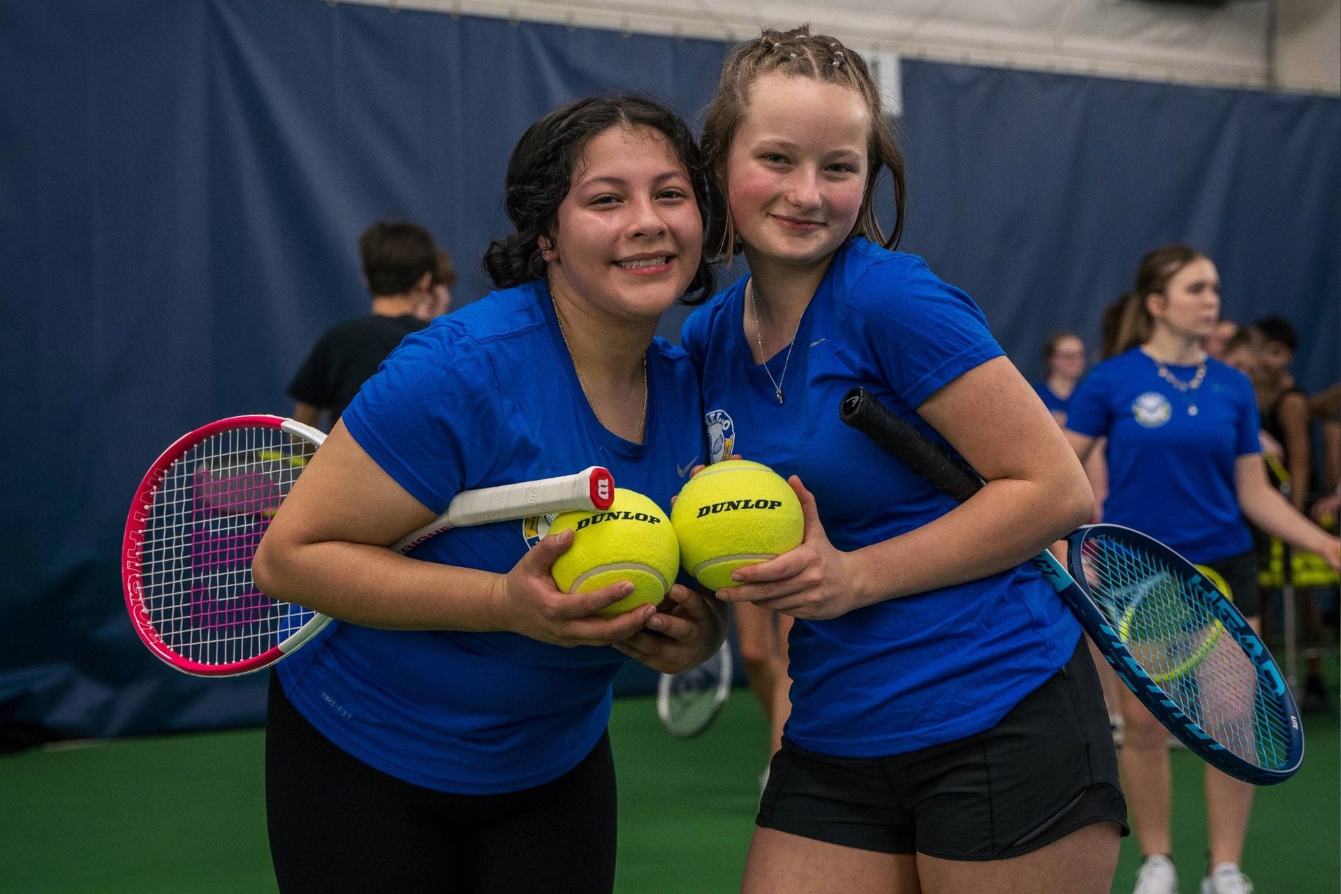 Two young women in blue shirts holding tennis balls and rackets, smiling at the camera, on an indoor tennis court.