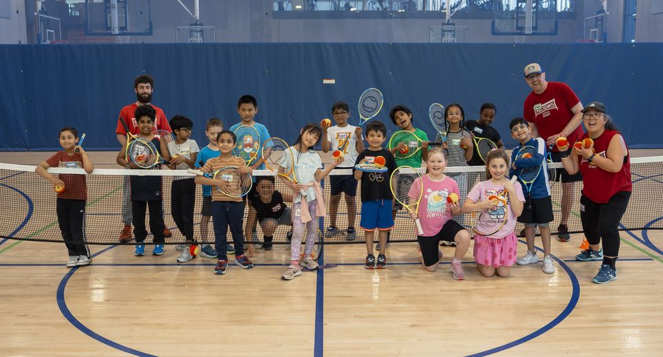 A group of children and two adults holding tennis racquets and balls, posing on an indoor court.