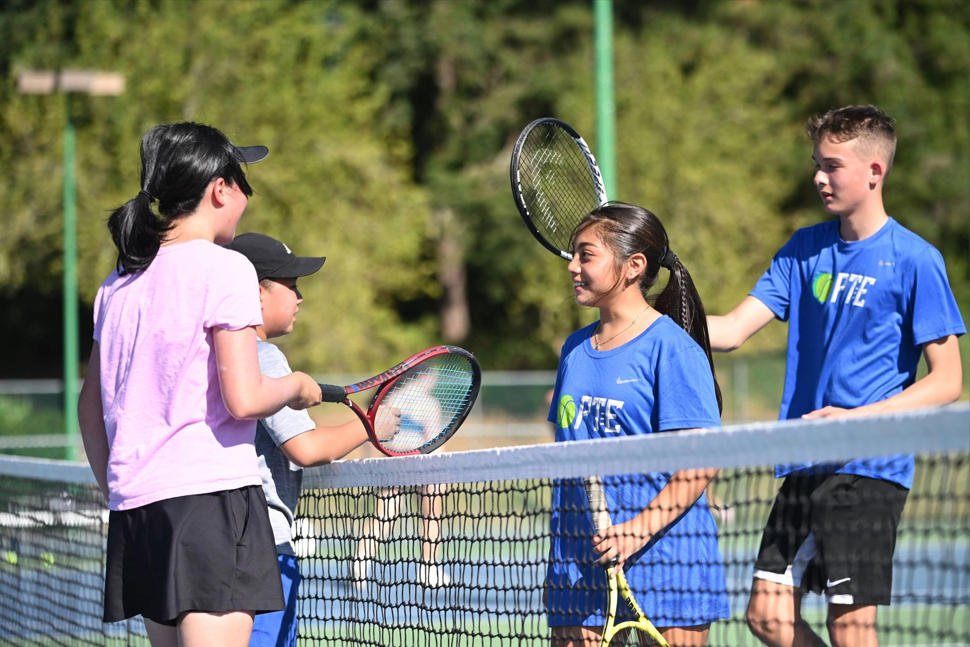 Four young tennis players shake hands over the net, all wearing sportswear, with a green, tree-lined background on a sunny day.