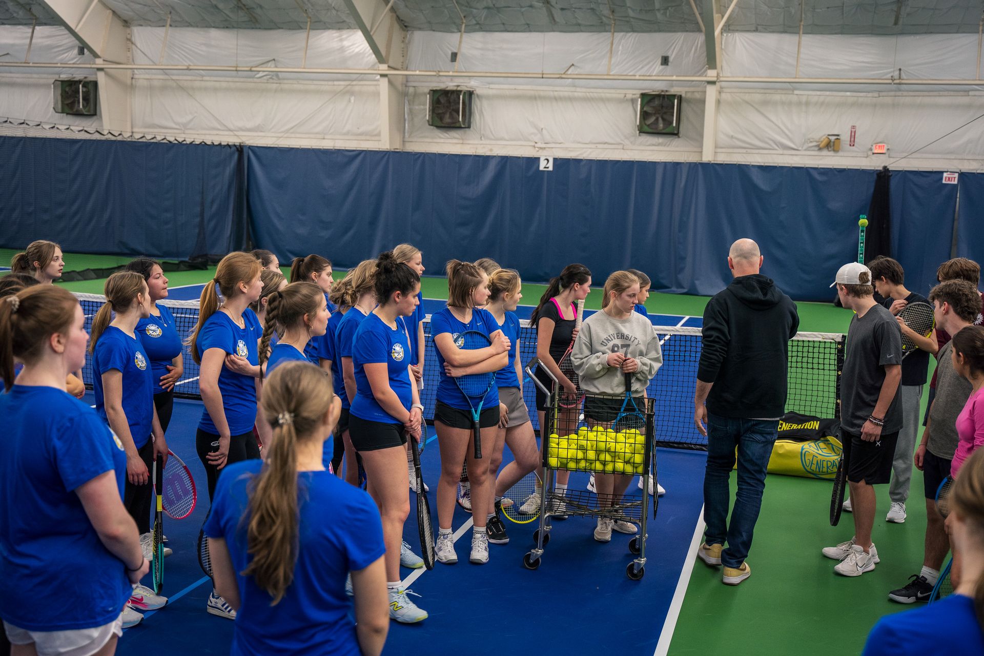 Indoor tennis practice with a coach explaining to a group of players in blue shirts, some holding rackets, near a cart of tennis balls.