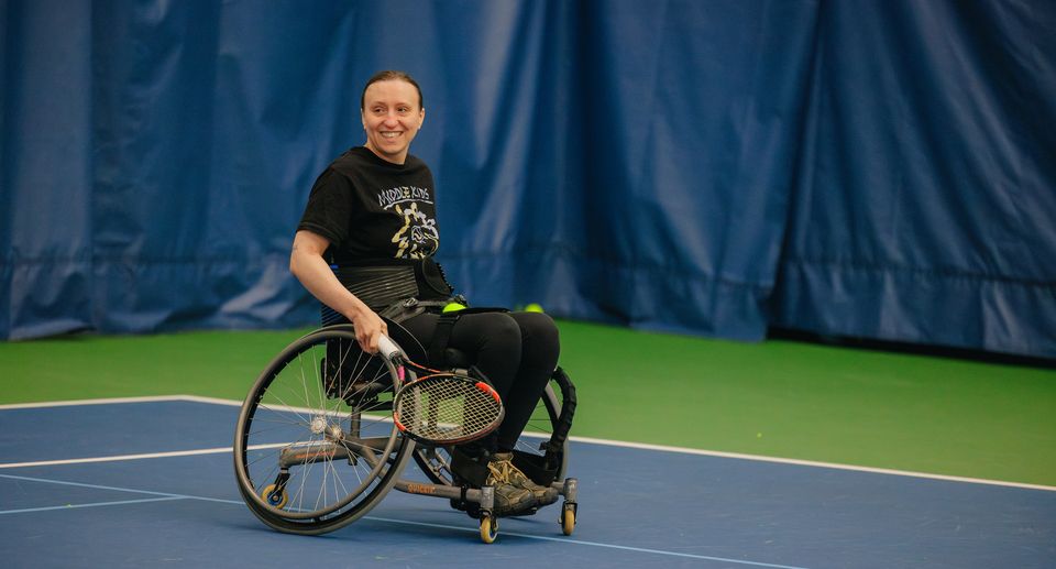 A person in a wheelchair holds a tennis racket on an indoor court, smiling and wearing a black T-shirt.