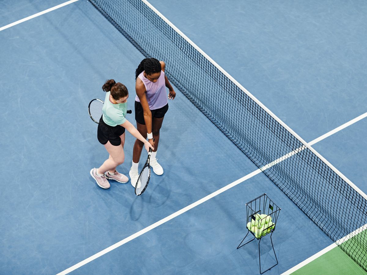 Two players stand on a tennis court, one adjusting the other's racket, with a basket of tennis balls nearby.