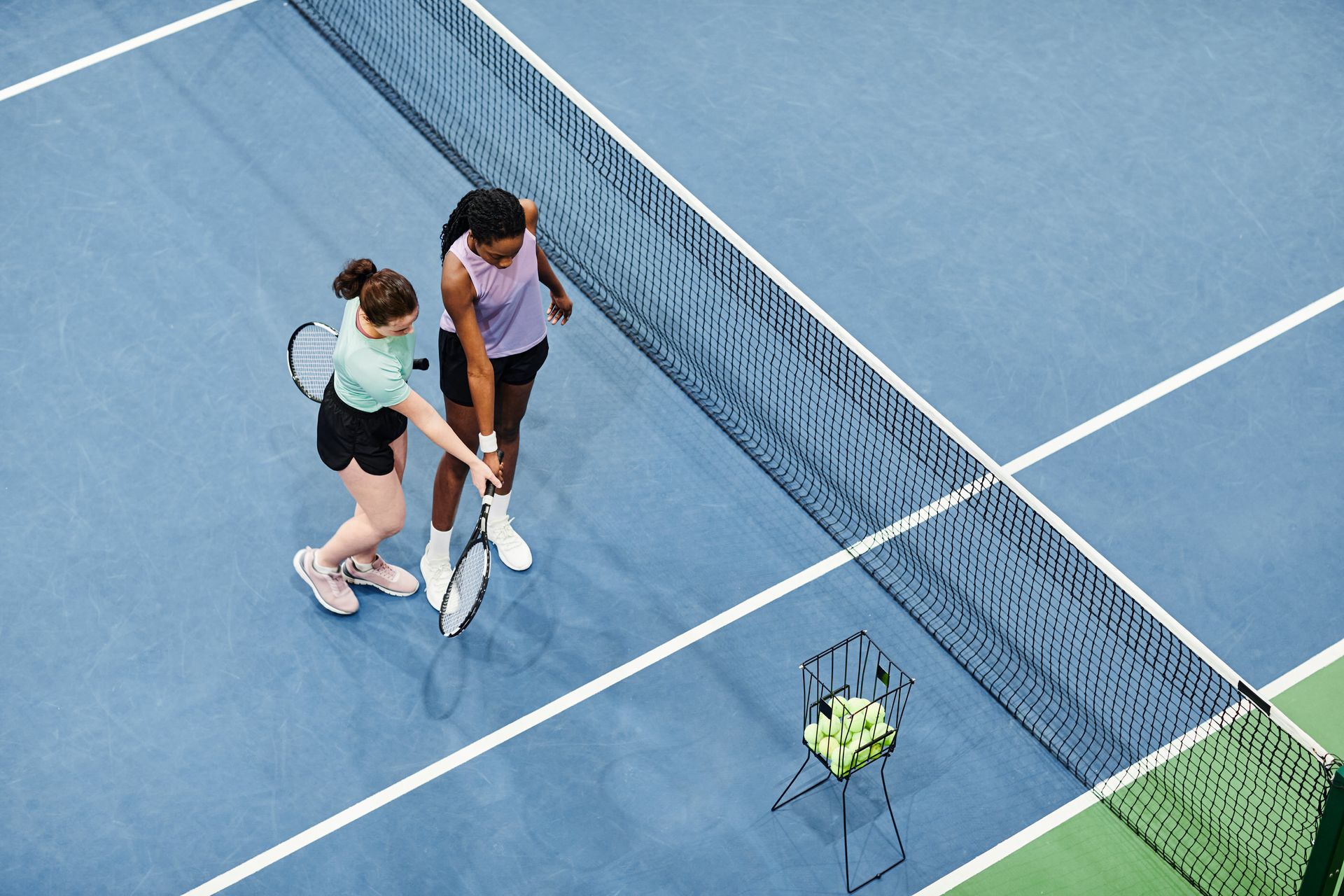 Two players stand on a tennis court, one adjusting the other's racket, with a basket of tennis balls nearby.