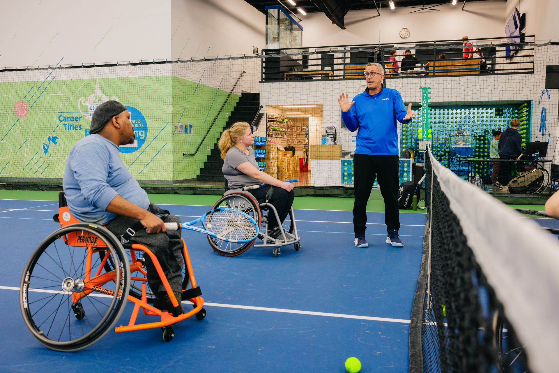Wheelchair tennis players listen to a coach on an indoor court, with sports equipment in the background.