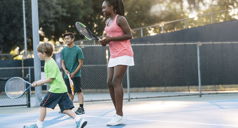 Three people enjoying a casual game of tennis on an outdoor court, with sunlight filtering through the trees.