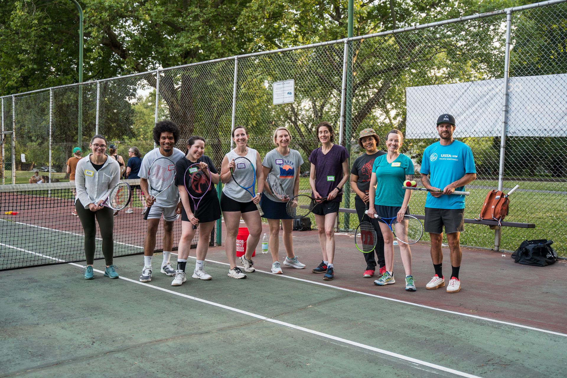 Group of people smiling and holding tennis rackets on a court, with trees and a net in the background.