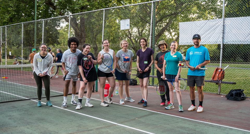Group of people smiling and holding tennis rackets on a court, with trees and a net in the background.