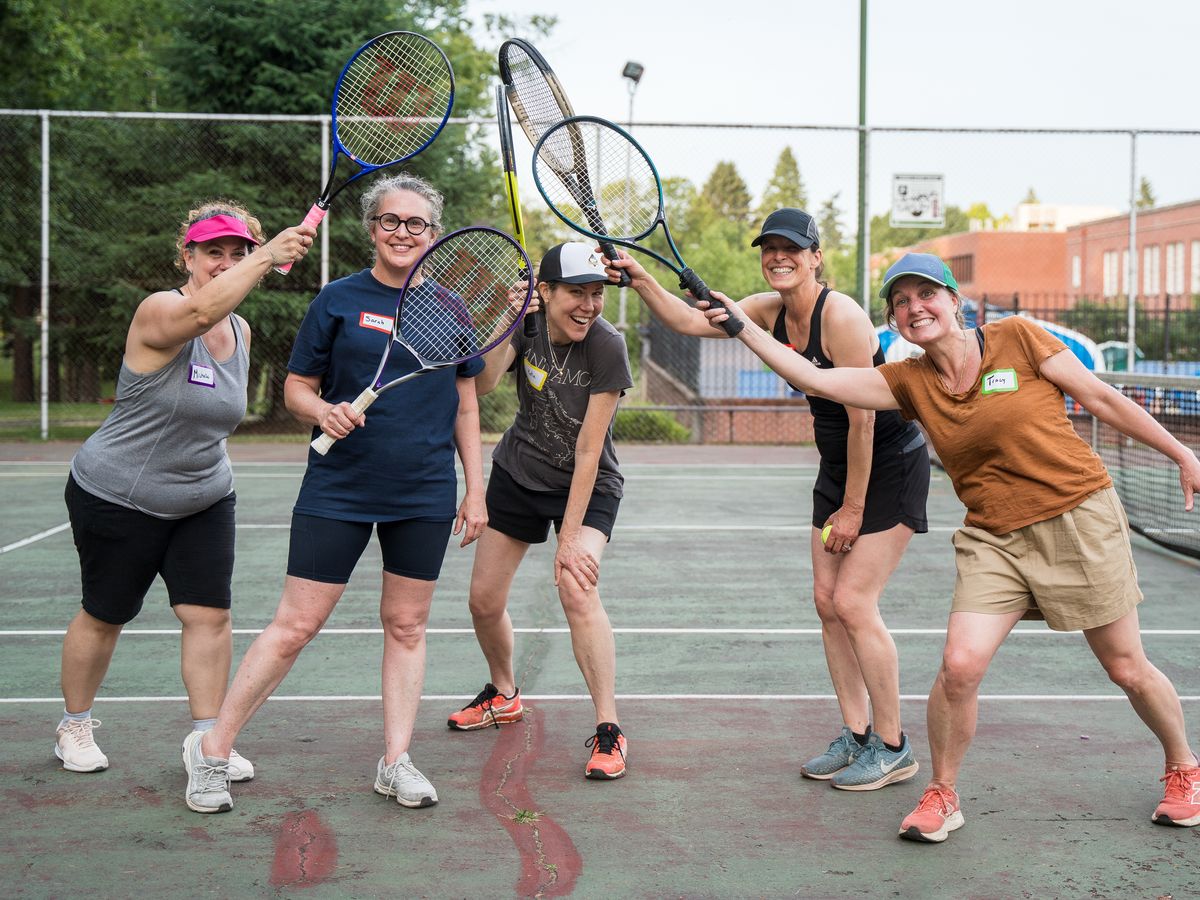 Five women smiling and holding tennis rackets, standing on an outdoor court with a background of trees and buildings.