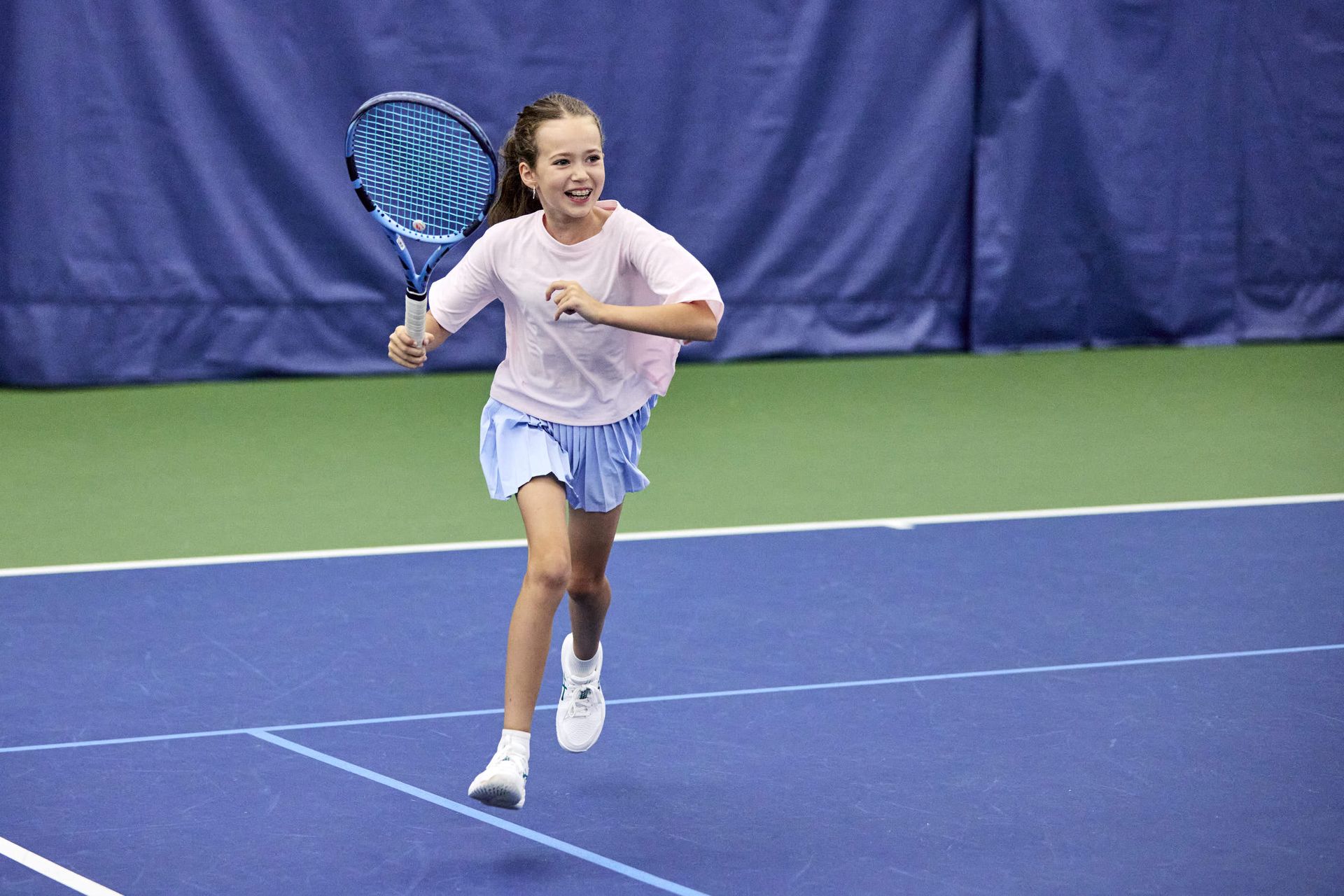 A young girl playing tennis indoors, running with a racket on a blue court, smiling and wearing a pink shirt and blue skirt.