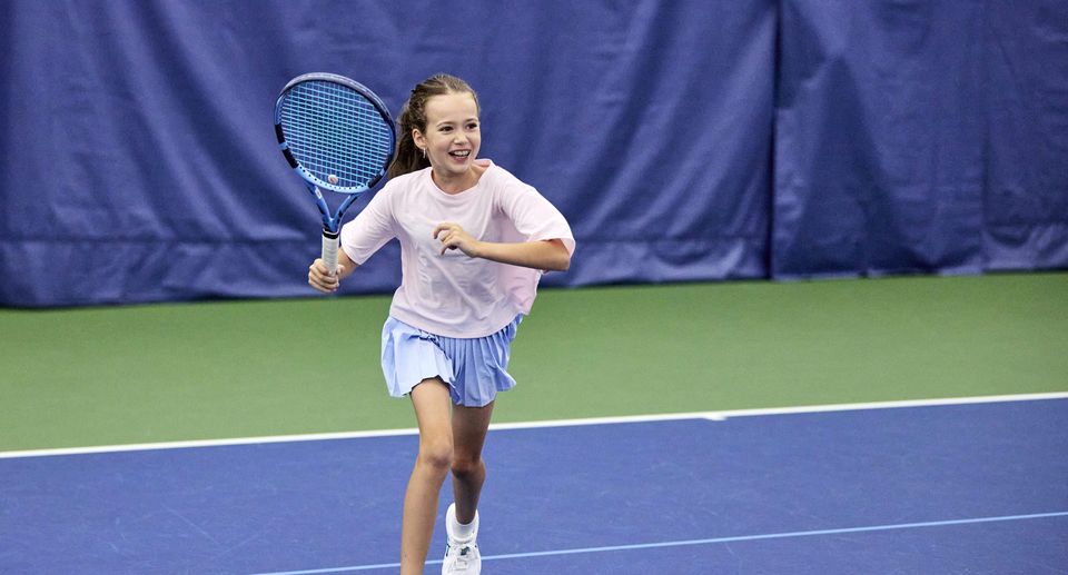 A young girl playing tennis indoors, running with a racket on a blue court, smiling and wearing a pink shirt and blue skirt.
