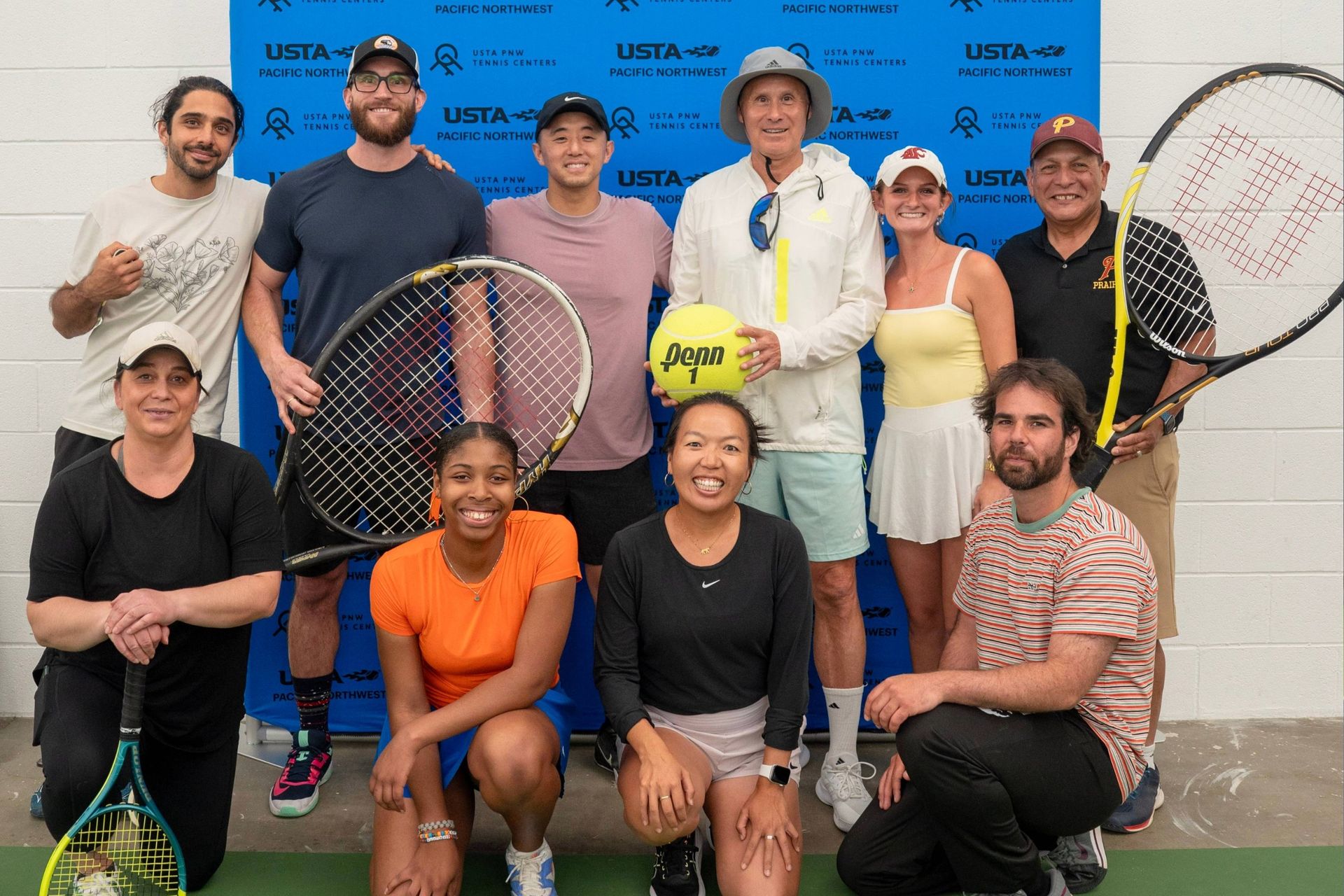 Group of ten people posing with tennis rackets and a large ball in front of a blue USTA backdrop, smiling, and dressed in athletic attire.