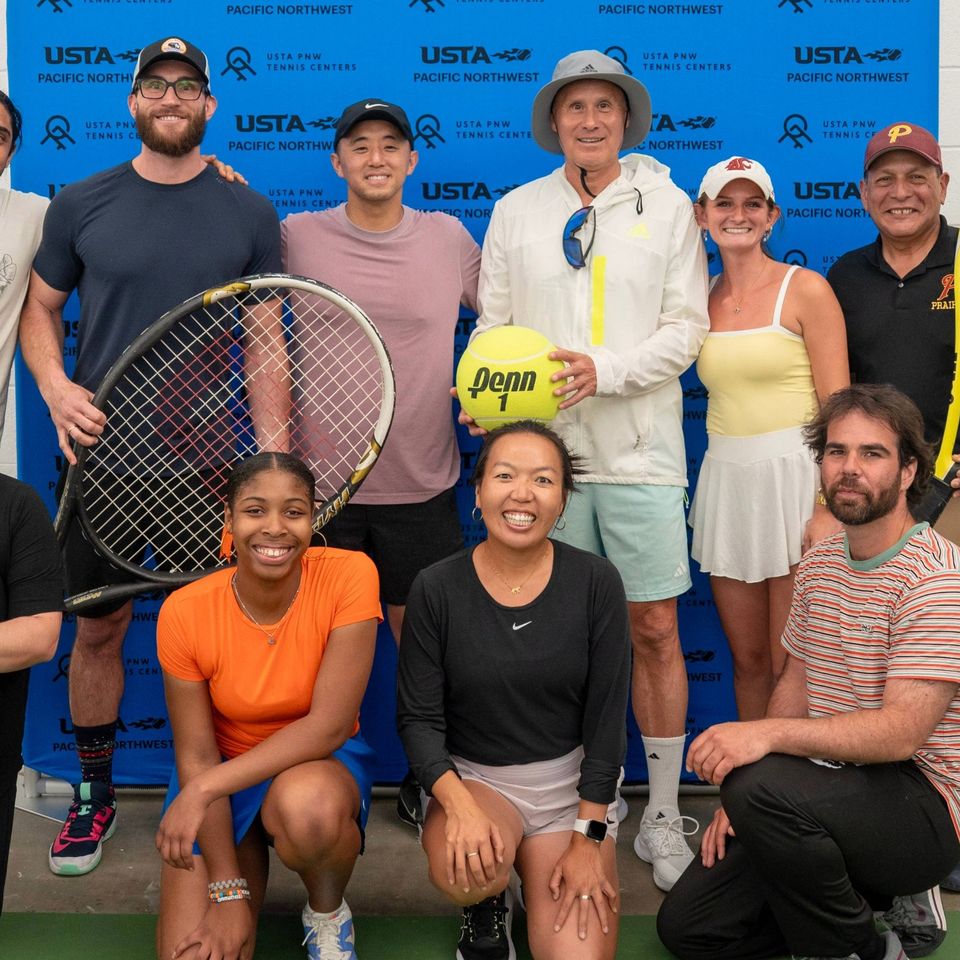 Group of ten people posing with tennis rackets and a large ball in front of a blue USTA backdrop, smiling, and dressed in athletic attire.
