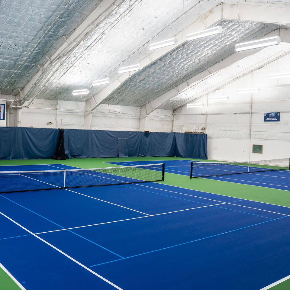 Indoor tennis courts with blue playing surfaces and green edges, under a high, white ceiling with bright lighting.
