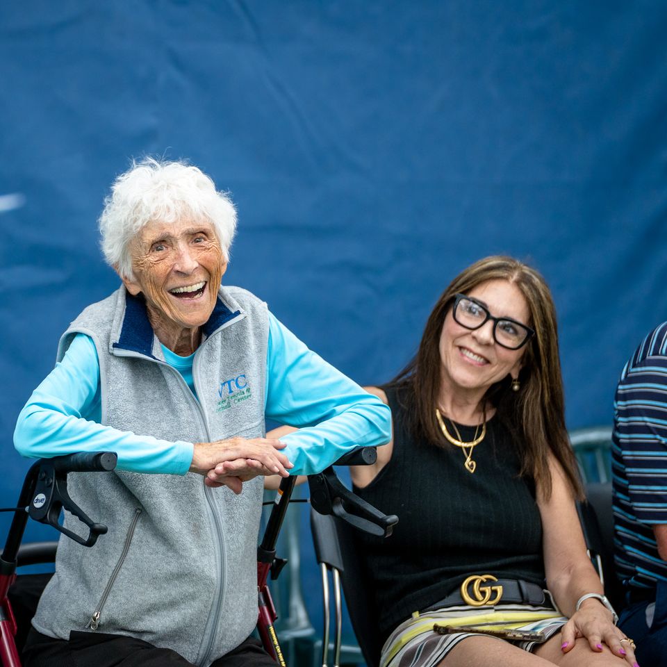 Elderly woman smiling, seated with a walker, next to a woman in glasses, listening to a person holding papers. Blue background.