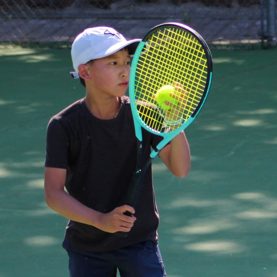 Young boy in a black shirt and white cap, playing tennis on a green court, focused on balancing a ball on his racket.