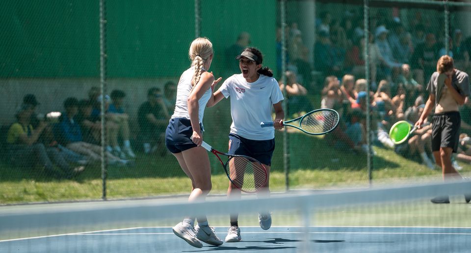 Two female tennis players celebrate on a court, high-fiving with rackets, while a crowd watches from behind a fence.