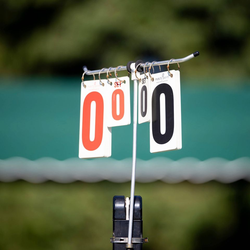 Close-up of a tennis score card showing two zeros in red and black against a blurred green background.