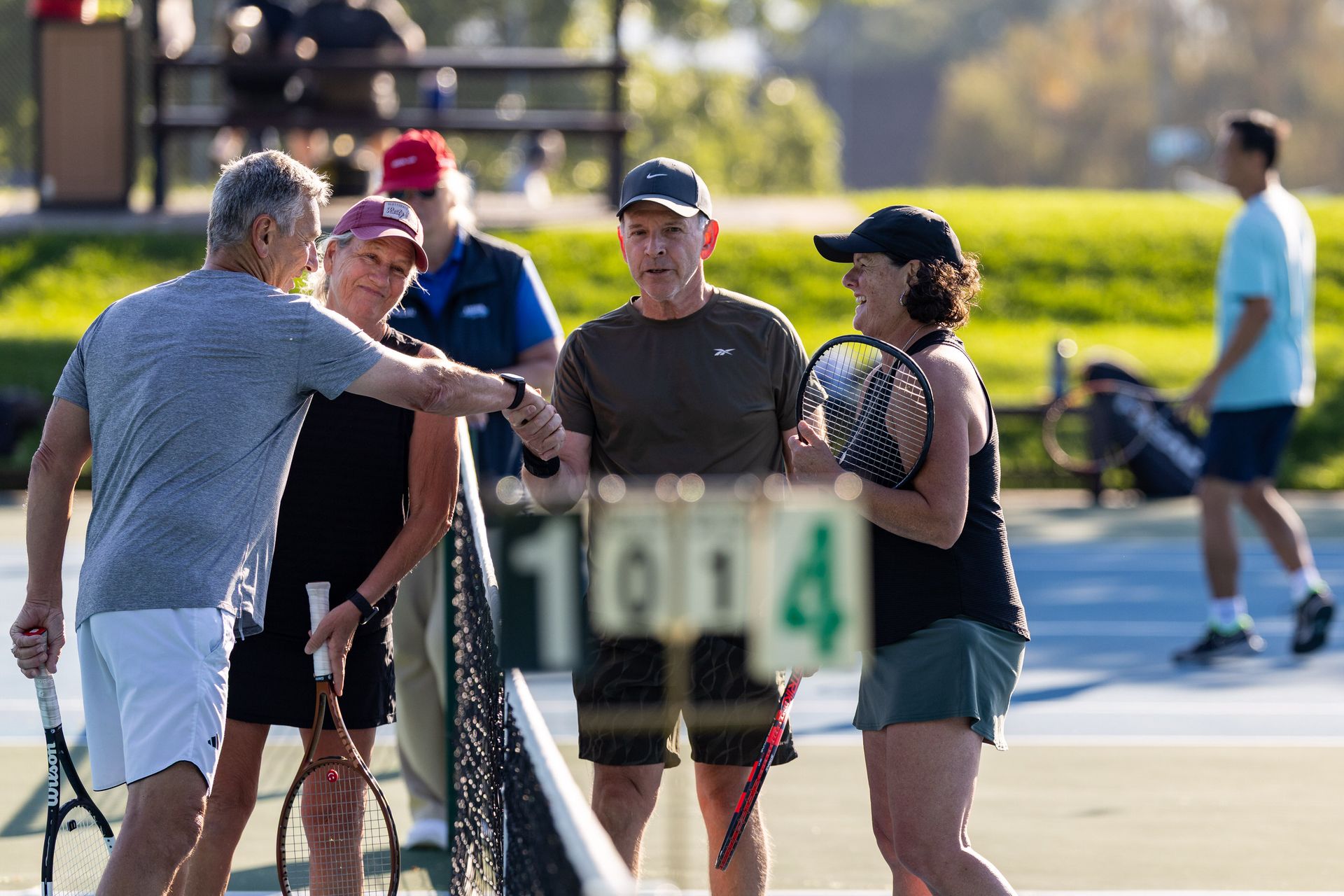 Four people with tennis rackets shake hands at the net after a match, smiling in sunlight. Scoreboard shows "1-0-4."