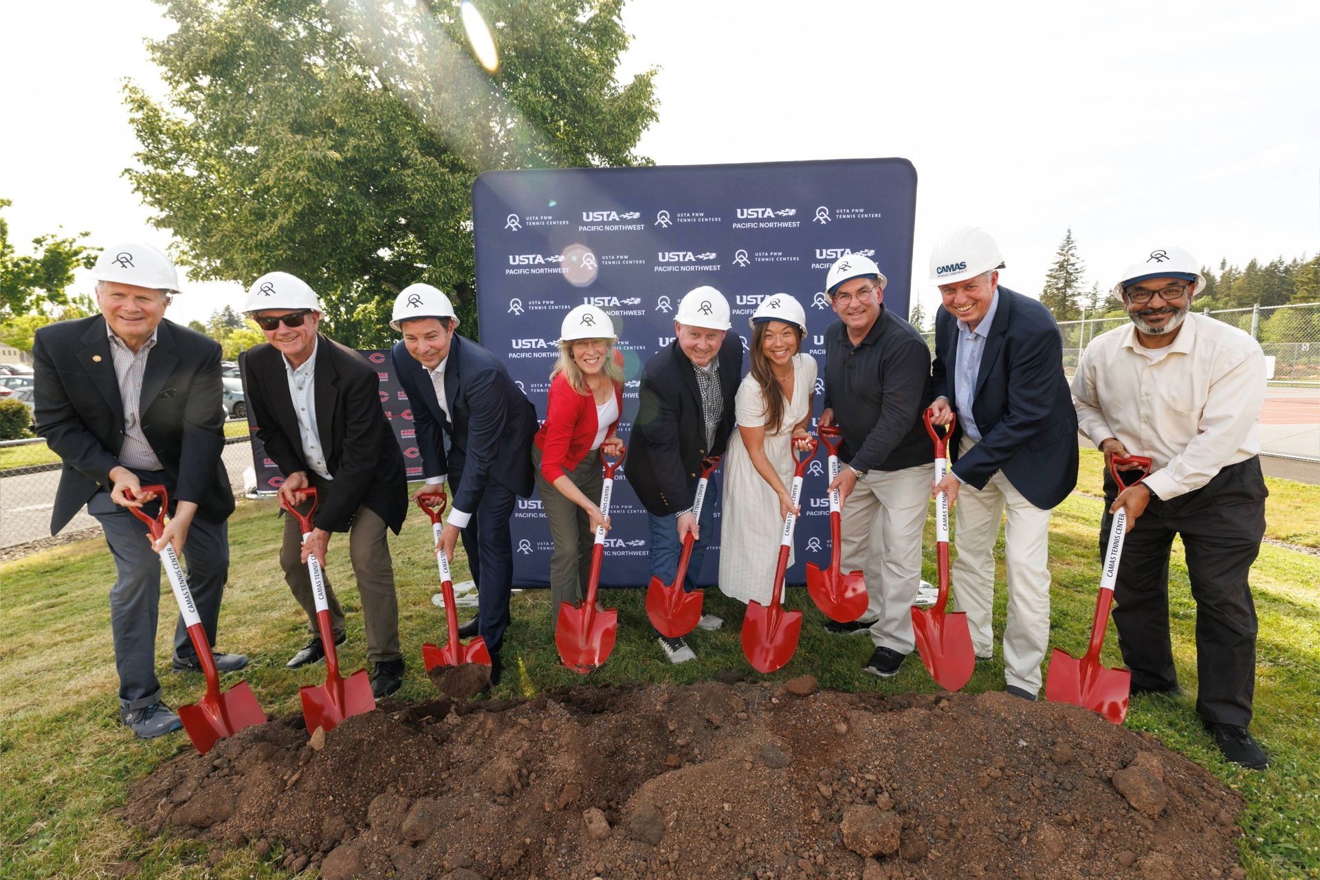 Nine people in hard hats holding red shovels during a groundbreaking ceremony, standing in front of a sign outdoors.